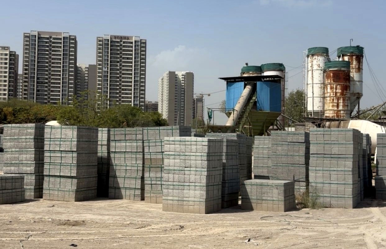 Stacked paver blocks at the Rewari supply site, with a batching plant and Gurugram skyline in the background